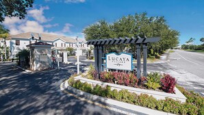 Un panneau indiquant l’entrée du complexe Lucaya Village, qui est sécurisée par une grille et agrémentée d’un espace paysager avec différentes espèces de plantes.