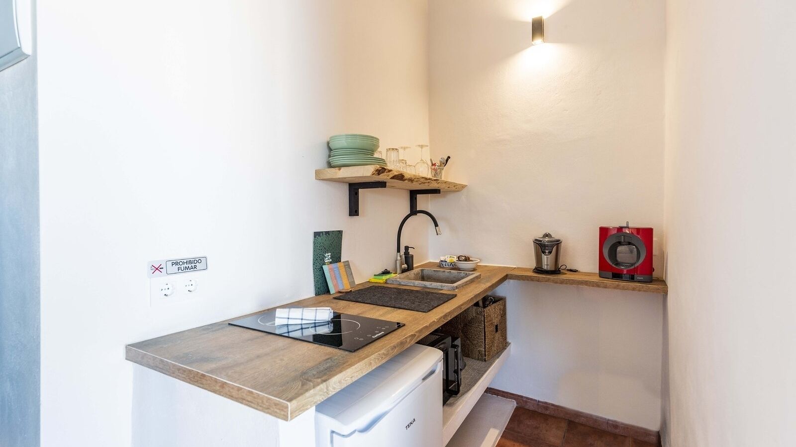 A compact kitchen with a built-in stove, a sink, and a coffee machine. There are shelves above the counter holding dishes and a small plant.