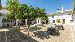 Une cour avec un sol carrelé, des sièges extérieurs et un bâtiment blanc orné d’arches et surmonté d’un toit en tuiles.