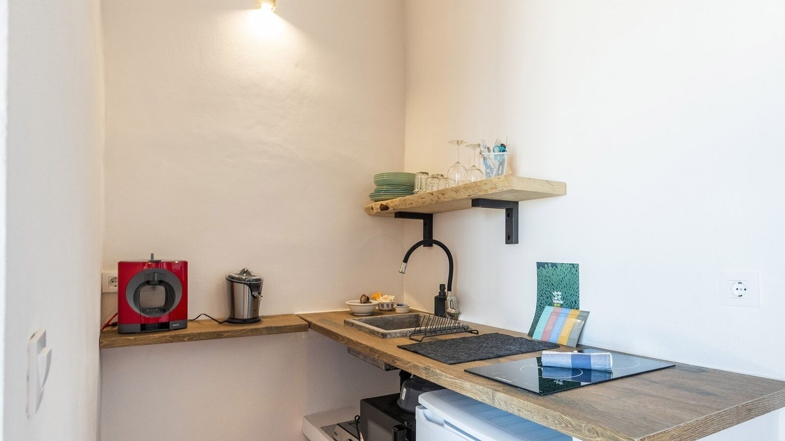 A compact kitchen with a coffee machine, a sink, a small shelf with glassware, and a book on the counter.