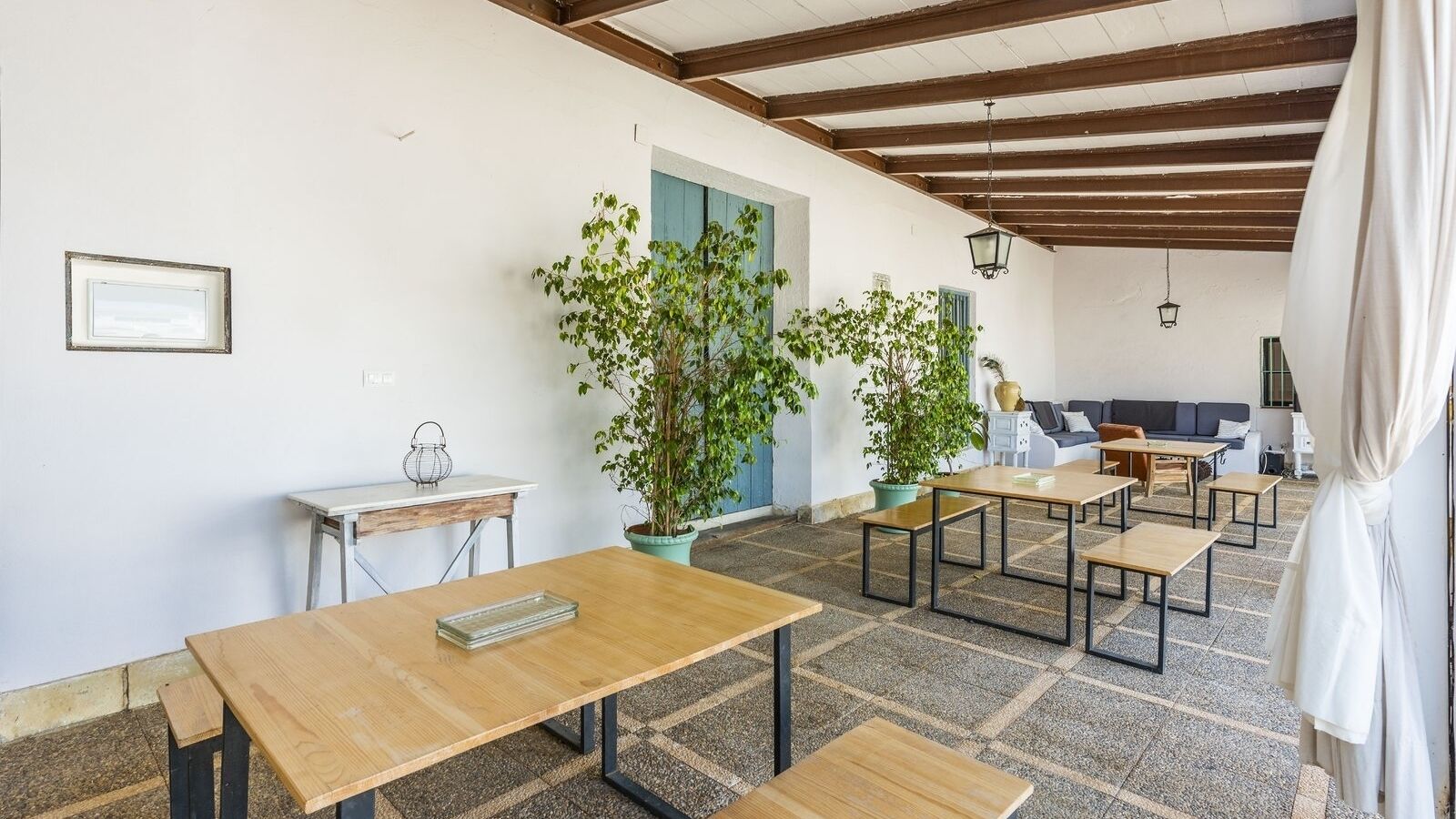 A spacious outdoor area with multiple wooden tables and benches, surrounded by potted plants and a blue door.