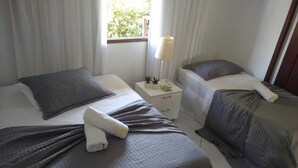 A hotel room with two beds, each with a grey quilted bedspread and pillows. There is a small bedside table with a lamp and a plant. - Flat no Condomínio Brisas do Amor (RN)