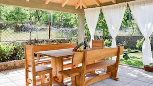 Une table et des bancs en bois, sous un toit en bois, avec un mur de pierre et de la verdure en arrière-plan.
