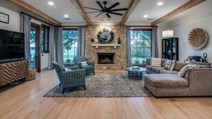 A spacious living room with a stone fireplace, a ceiling fan, and a large TV. - Luxury Lakefront Estate - 1.6 Acres - Sleeps 12 - Boathouse + Sunset Views (Little Elm)