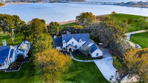 A large, white house with a stone entrance, surrounded by well-maintained lawns and trees, near a body of water with a bridge in the background. - Luxury Lakefront Estate - 1.6 Acres - Sleeps 12 - Boathouse + Sunset Views (Little Elm)