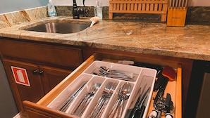 Una cocina con encimera de granito, pileta de acero inoxidable y organizador de cajones con utensilios.