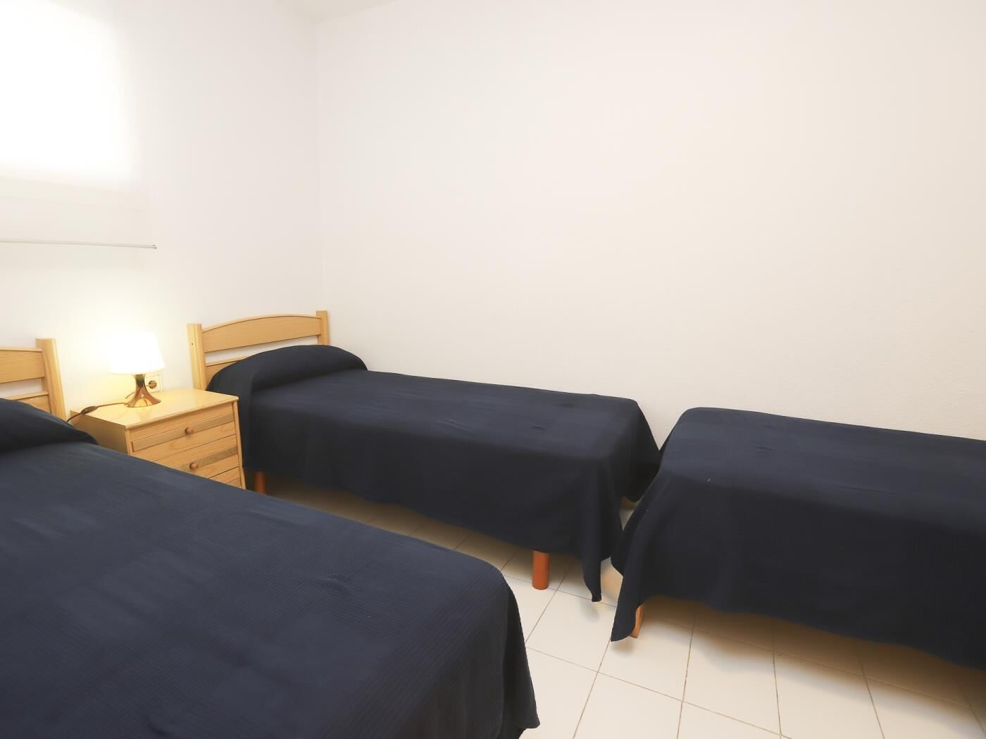Habitació amb tres llits individuals, una taula de nit de fusta amb una làmpada i una paret blanca sense decoració.