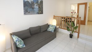 A living room with a sofa, a potted plant, a wooden railing, and a kitchen in the background.