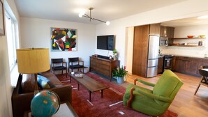 Una sala de estar moderna con un sofá, una mesa de centro y un sillón verde. Un globo y una lámpara están sobre el suelo. Se ve una cocina con un refrigerador y microondas al fondo.