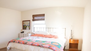 Un dormitorio con cama, mesita de noche, lámpara, silla y un cuadro en la pared.
