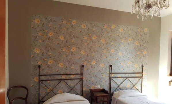 Une chambre avec deux lits, une chaise en bois, une table de chevet et un papier peint à motifs floraux.