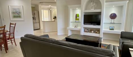 A living room with a gray sofa, a coffee table, and a TV on a white wall. There is a dining area with a wooden table and chairs.