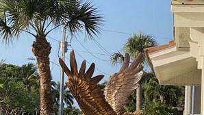 A bronze eagle statue on a pedestal in a landscaped garden with palm trees and a house in the background. - EASY 5-Min Walk to Beach Path! NEW Saltwater Pool & Spa, Steps to Dining & Shops (Marco Island)