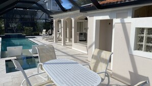 An outdoor pool area with lounge chairs and a table, surrounded by a covered patio and a building with columns. - EASY 5-Min Walk to Beach Path! NEW Saltwater Pool & Spa, Steps to Dining & Shops (Marco Island)