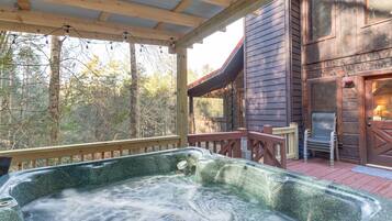 Un bain à remous installé sous une terrasse en bois, avec vue sur un bâtiment rustique et des arbres.
