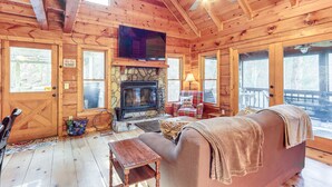 Interior acolhedor de uma cabana, com lareira de pedra, paredes de madeira, televisão e ventilador de teto.