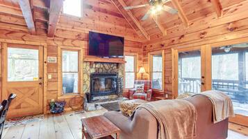 L’intérieur chaleureux d’un chalet avec une cheminée en pierre, des murs en bois, un téléviseur et un ventilateur de plafond.