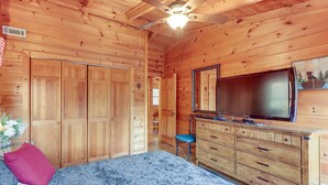 Quarto de cabana de madeira com cama, penteadeira, televisão e ventoinha no teto.