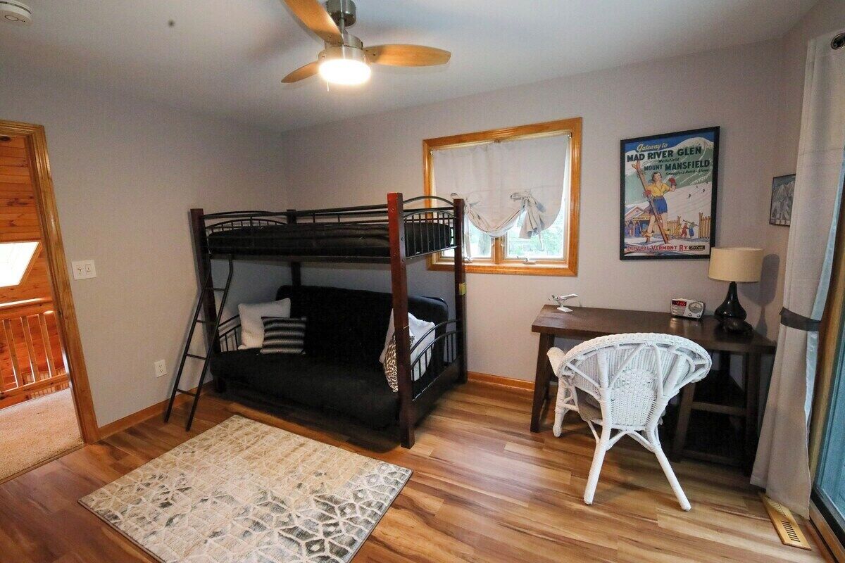 Une chambre avec un lit superposé, un bureau avec une chaise, une fenêtre avec des rideaux, une affiche au mur et un ventilateur de plafond.