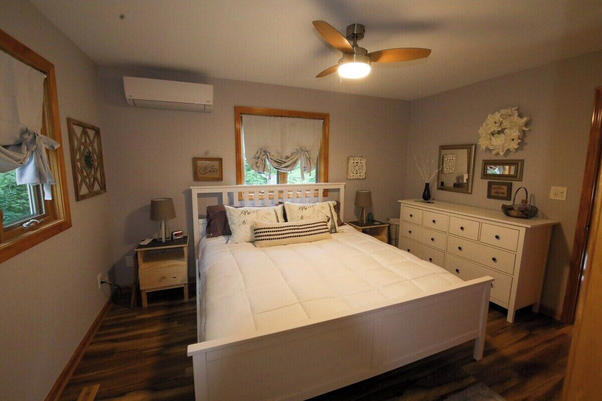 Un dormitorio con cama, mesita de noche, cómoda, ventilador de techo y ventana con cortinas.