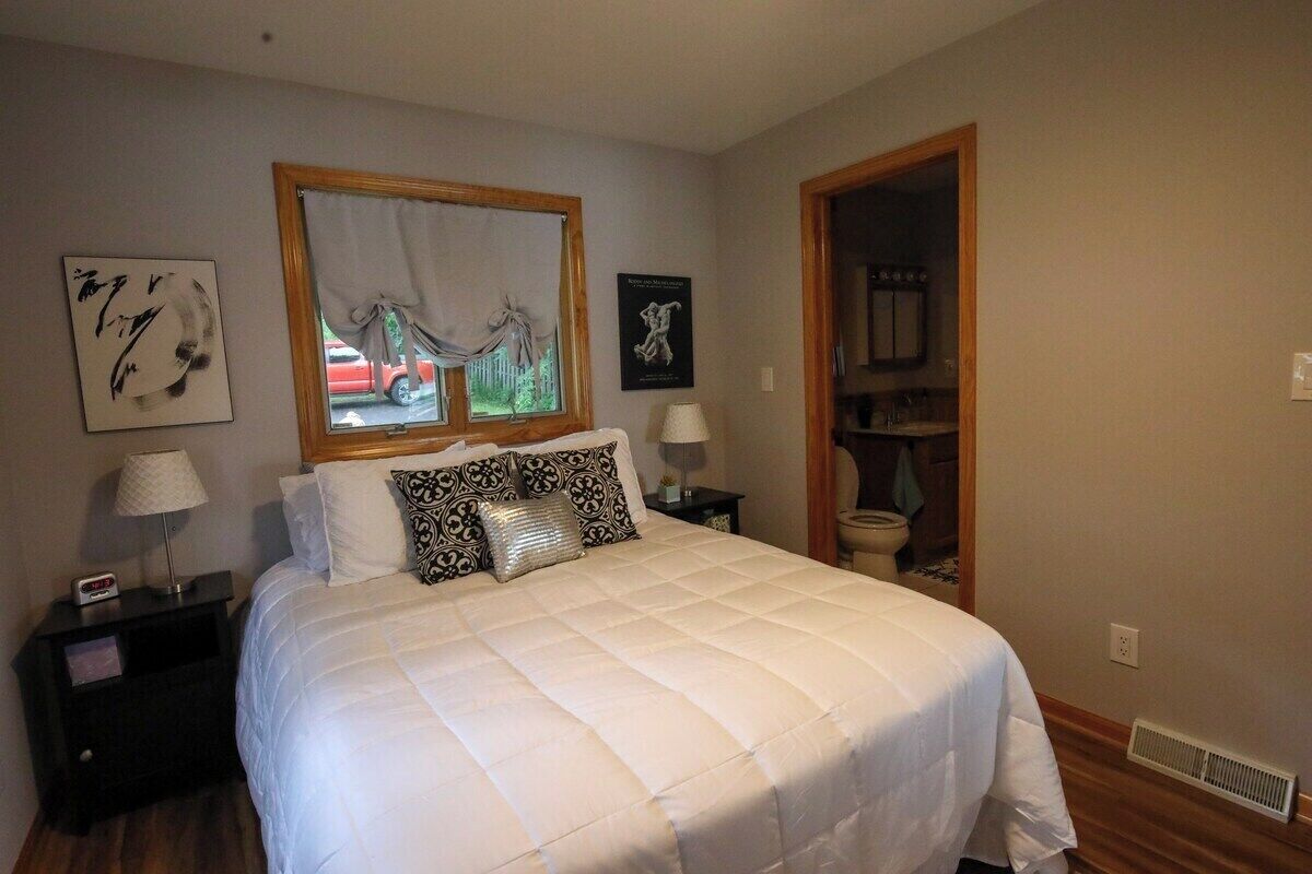 Una cama bien hecha con ropa blanca y almohadillas decorativas, una mesita de noche con lámpara, una obra de arte enmarcada sobre la ventana, y una puerta de madera que conduce a un baño.