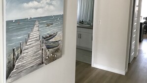 A hallway with a painting of boats on a pier, a kitchen area, and a door leading to another room.