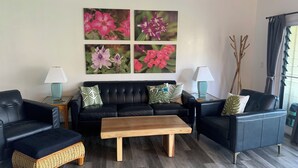 Een woonkamer met een zwarte leren bank, twee bijzettafels met lampen, een houten salontafel, en een muur met vier ingelijste bloemschilderijen.