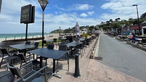 An outdoor café with tables and chairs, a signboard, and a view of the beach and ocean. - Appartement Résidence le Casino à Perros Guirec (PERROS GUIREC)