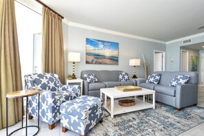 Una sala de estar con un sofá gris, un sillón a juego y un reposapiés, una mesa de centro blanca y un cuadro grande de una escena de playa.