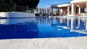 A swimming pool with clear blue water, surrounded by a white pool deck and a covered area with lounge chairs.