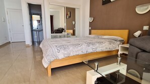 A modern bedroom with a bed, a sofa, a glass coffee table, and a bathroom visible through an open door.