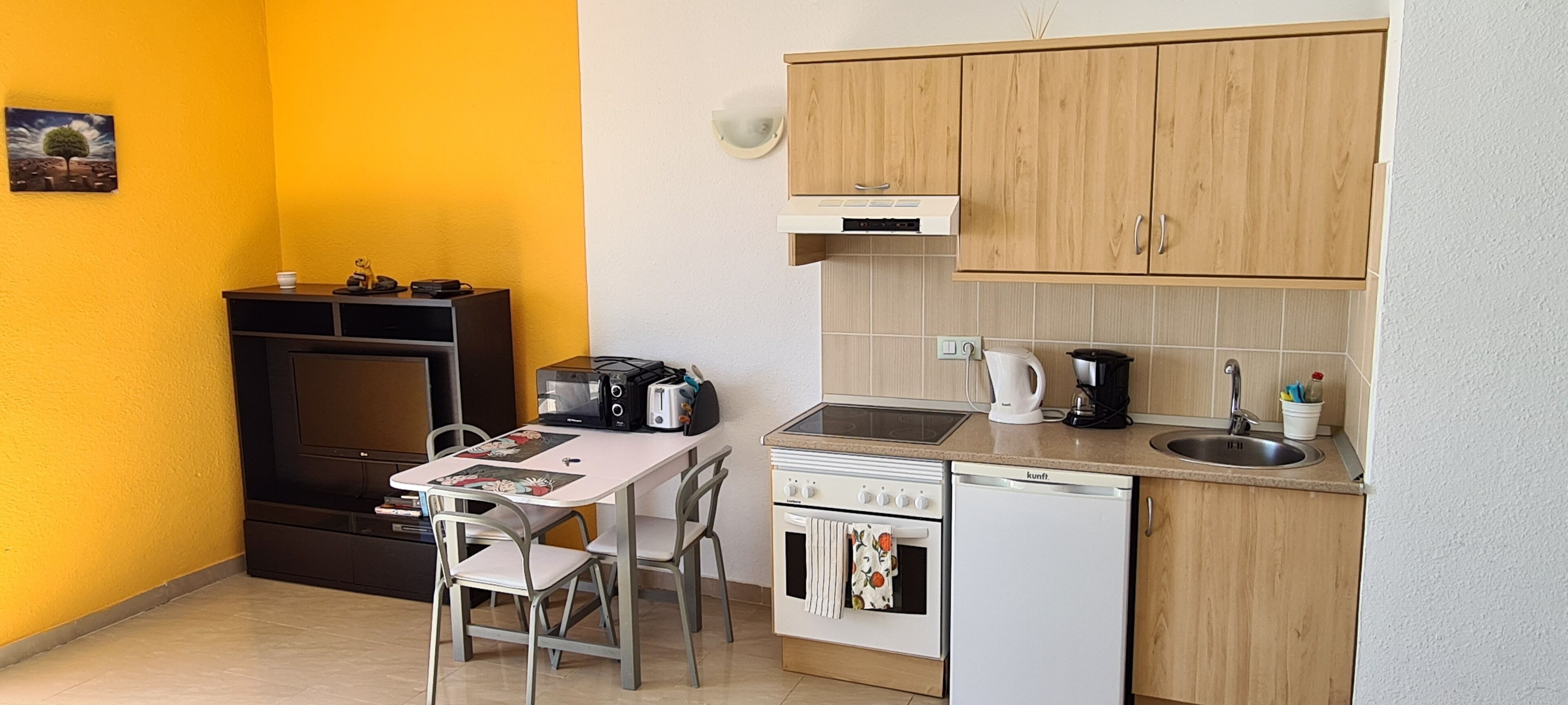 Una cucina compatta con piano cottura, forno e lavello, un piccolo tavolo da pranzo con sedie e una parete gialla.
