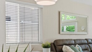 Ein modernes Wohnzimmer mit einer braunen Ledersofa, einem weißen Tisch mit Pflanze und einem großen Fenster mit Jalousien.
