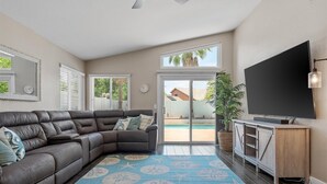 Ein Wohnzimmer mit einem Ecksofa, einem Flachbildfernseher, einer Holzkommode und einem großen Fenster mit Blick auf einen Pool und Palmen.