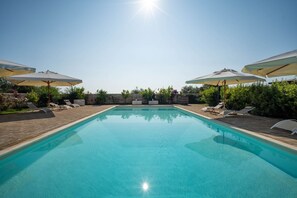 Una piscina azul clara con sillas de descanso y sombrillas, rodeada de follaje.
