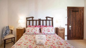 A bedroom with a floral bedspread, two bedside tables with lamps, a wooden door, and a chair with a blanket. - TERRA TERRA - Masseria Minunni - Suite C.Tartara (Conversano)