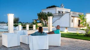 A couple relaxing on outdoor white wicker furniture with a view of a modern white building and a pool in the background. - TERRA TERRA - Masseria Minunni - Suite C.Tartara (Conversano)