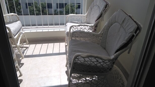 A balcony with two white wicker chairs and a small table, overlooking a building with a white facade and a metal roof.