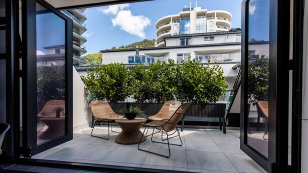 En balkon med to fletstole og et rundt bord, med udsigt over en moderne bygning med balkoner og en klar himmel.