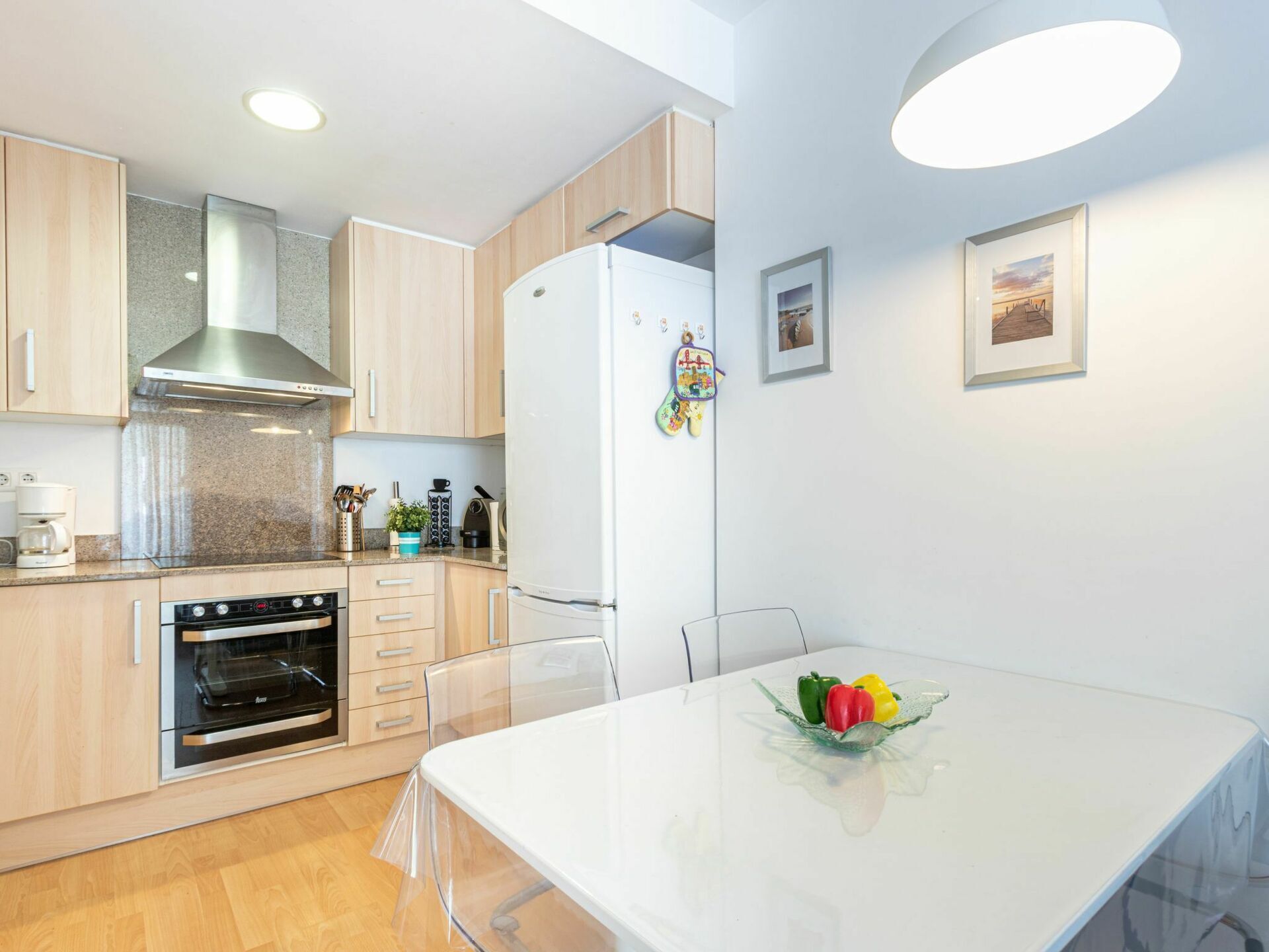 Una cocina moderna con armarios de madera, una campana extractora de acero inoxidable, horno empotrado y refrigerador blanco. Hay una mesa de comedor con cubierta de vidrio transparente y un cuenco de fruta.