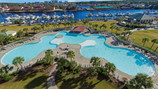 A large, irregularly shaped swimming pool with a small island in the center, surrounded by palm trees and a fence. - 🏝 Barefoot Landings in N Myrtle Beach-2 Resort Pools/ Golf/  8 Mins to Beach 🏝 (North Myrtle Beach)