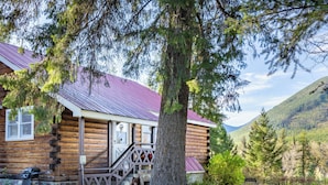 Une cabane rustique en rondins de bois, avec un toit en métal, une véranda et un grand arbre devant.