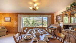 Une salle à manger avec une table en bois dressée pour le repas, un piano et une vue sur une montagne boisée.
