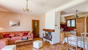 A cozy living room with a wooden floor, a pink sofa, a white ottoman, and a fireplace. There is a dining area with a table and chairs, and a kitchen with wooden cabinets and a window. - L'ensoleillade by Interhome (Saint Cyr sur mer Les Lecques)