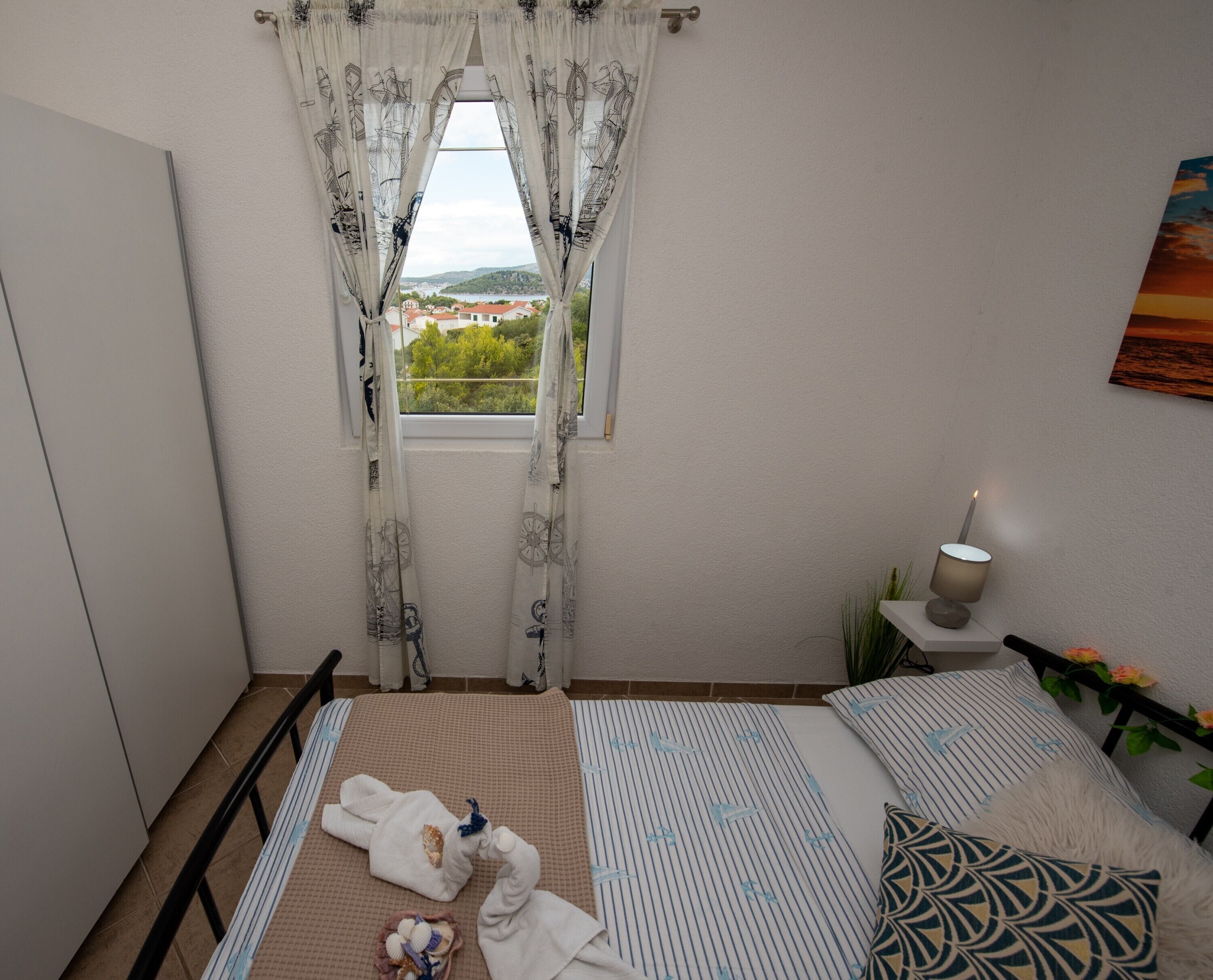 Une chambre à coucher avec un lit, une table de chevet avec une lampe, une fenêtre avec des rideaux et une vue sur la verdure à l’extérieur.