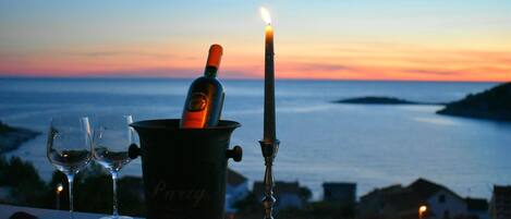 A candle, two wine glasses, and a bottle of wine on a balcony overlooking a body of water at sunset.