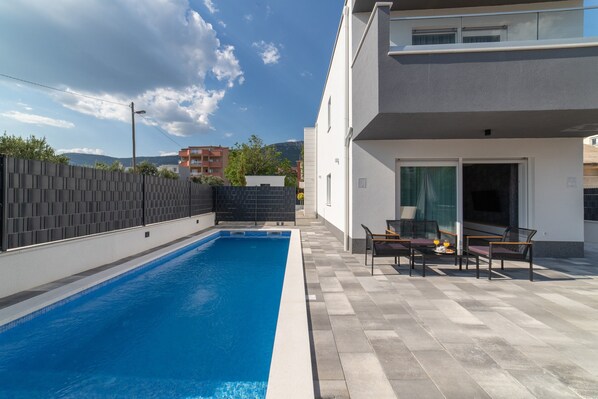 Un área de piscina exterior moderna con solárium empedrado, zona de estar con sillas y mesa, y vistas de edificios y colinas circundantes.