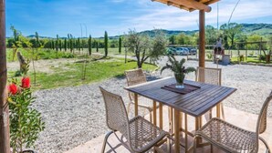 Pátio externo com mesa de madeira e cadeiras de vime, com vista para um vinhedo e um estacionamento.