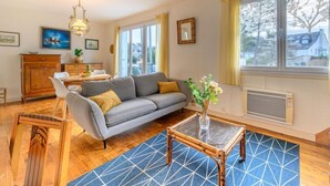 En stue med sofa, et sofabord i glas, spiseområde med bord og stole, samt store vinduer med gardiner.