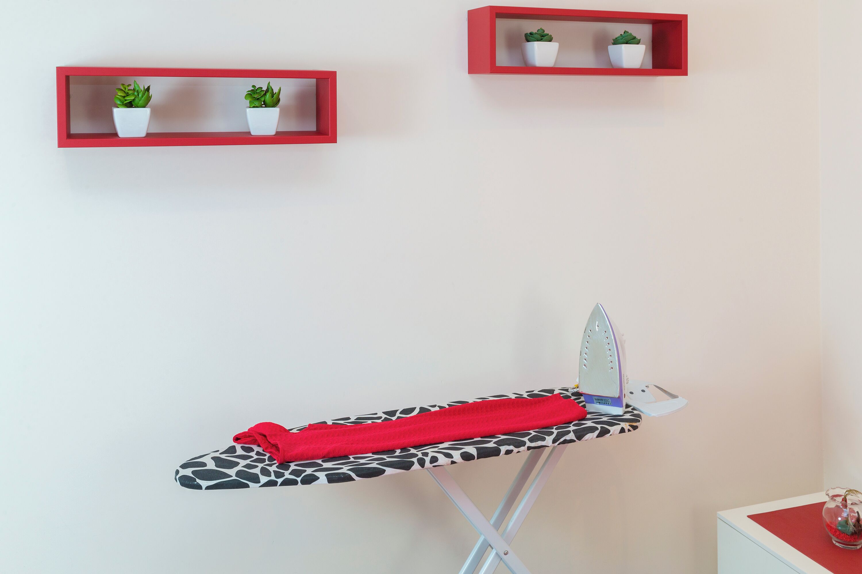 Una tabla de planchar con una toalla roja, un planchador blanco y una funda estampada con diseño de jirafa. Se observan dos repisas rojas con plantas en macetas.